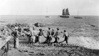 Een aanlegplaats op Saba, op het strand staat een een groep mannen die passagiers van en naar boten roeit, iets verder op zee ligt een zeilschip (Collectie Wereldmuseum, TM-10021355)
