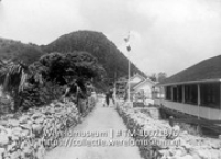 Gouvernementshuis op Saba (The Bottom); Een smalle straat met aan weerszijde stenen wallen en voor het gouvernementshuis een wapperende vlag in de top van de vlaggenmast (Collectie Wereldmuseum, TM-10021376)