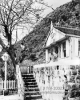 Huisje in The Bottom, Saba. Het wordt thans bewoond door de enige onderwijzer van Saba; Een man hangt over de balustrade van zijn woning (Collectie Wereldmuseum, TM-10021391)