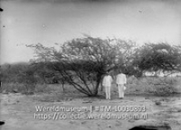 Twee mannen in uniform poseren bij een uitgedroogde boom op zandgrond (Collectie Wereldmuseum, TM-10030893)