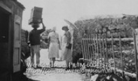Saba het dragen van lasten op het hoofd; Smal pad met rechts een hek en links bebouwing waarop twee vrouwen in zomerjurken een man passeren die een kist op zijn hoofd draagt (Collectie Wereldmuseum, TM-60047578)