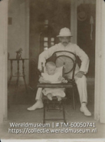 Baby Wilhelm de Gaay Fortman in een kinderstoel met daarachter zijn vader op een omgekerde stoel in een wit pak met tropenhelm (Collectie Wereldmuseum, TM-60050741)